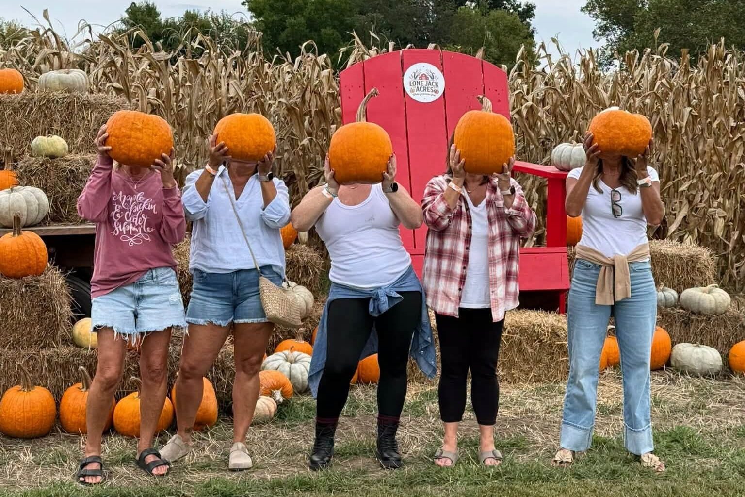 Five guests holding pumpkins in front of their faces by the giant red Lone Jack Acres chair, with hay bales, pumpkins and corn stalks behind them